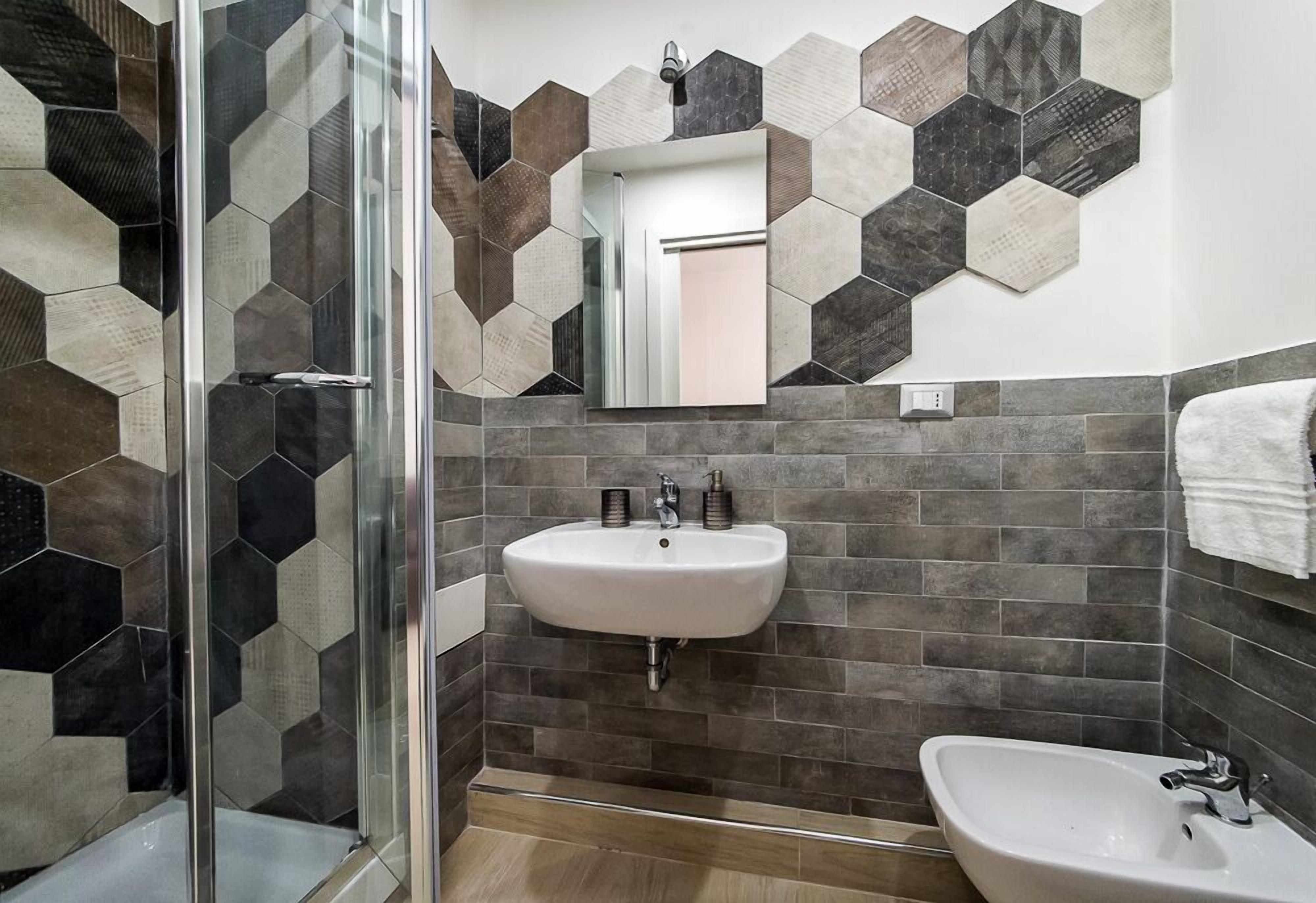 A bathroom with a glass shower enclosure, a sink with a mirror above it, and a tiled wall with hexagonal patterns.