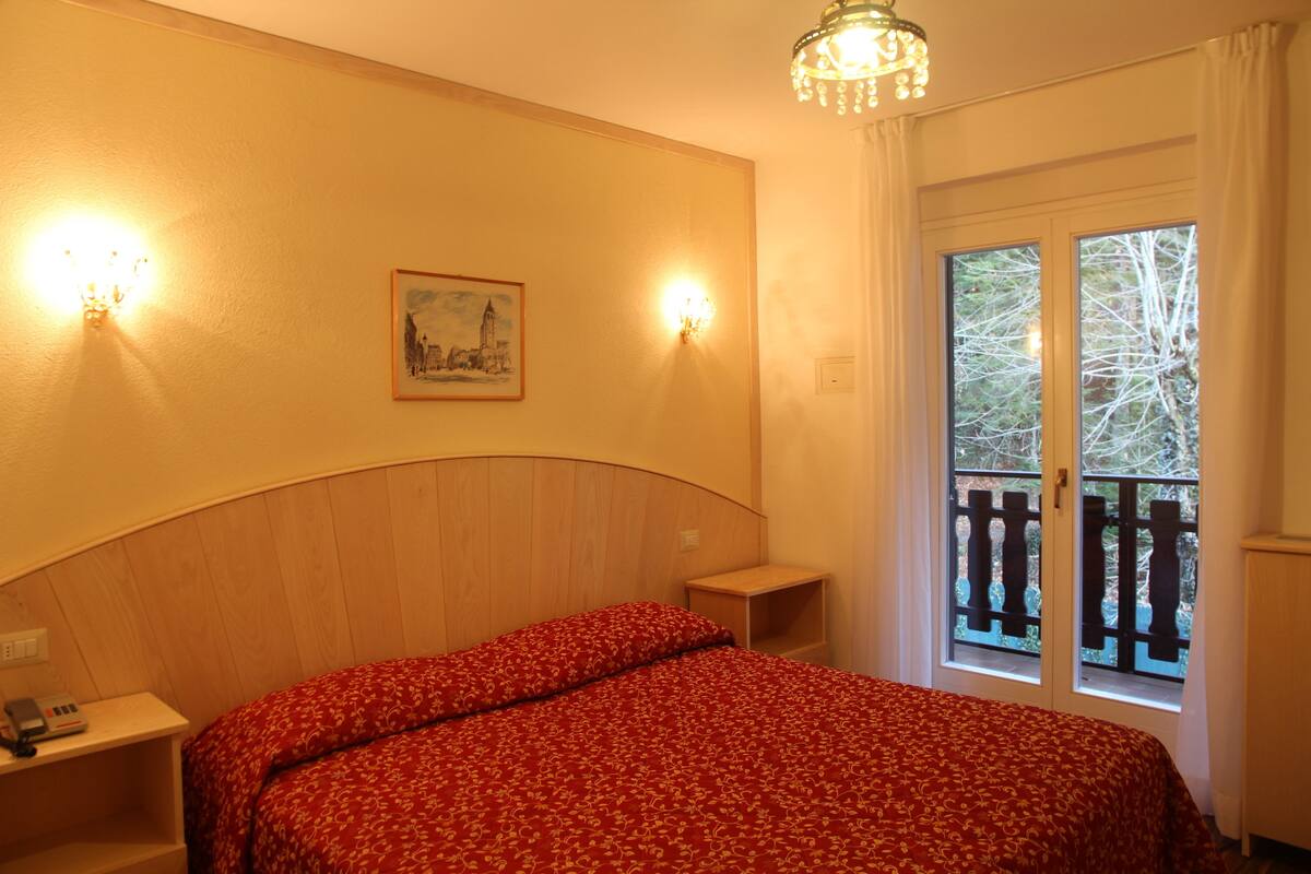 Un dormitorio con cabecera de madera, colcha floral roja, dos apliques de pared, una mesita de noche con un teléfono y una ventana con vistas a árboles.