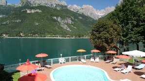 Piscina com espreguiçadeiras e guarda-sóis, cercada por montanhas e um lago.