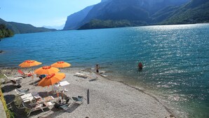 Uma praia com guarda-sóis e espreguiçadeiras, um lago calmo e montanhas ao fundo.