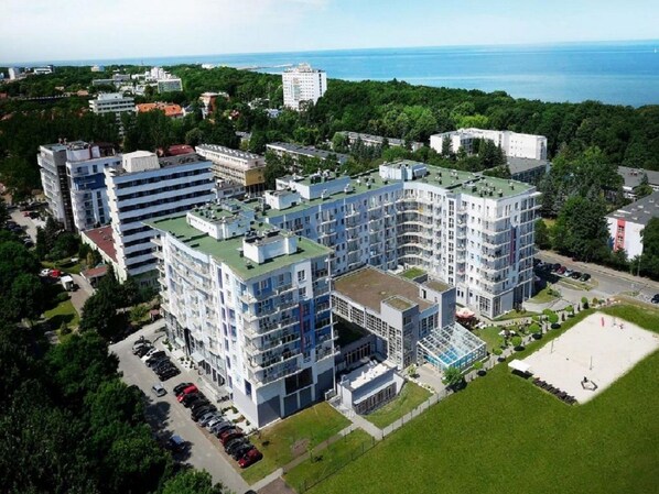 Un immeuble moderne à plusieurs étages, doté d’une terrasse sur le toit, entouré de verdure et comprenant un espace de stationnement.