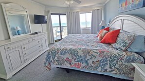 A bedroom with a bed, a dresser, a television, and a balcony with a view of the ocean.