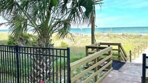 A wooden pathway leading to a beach with a metal fence and potted plants with pink flowers.