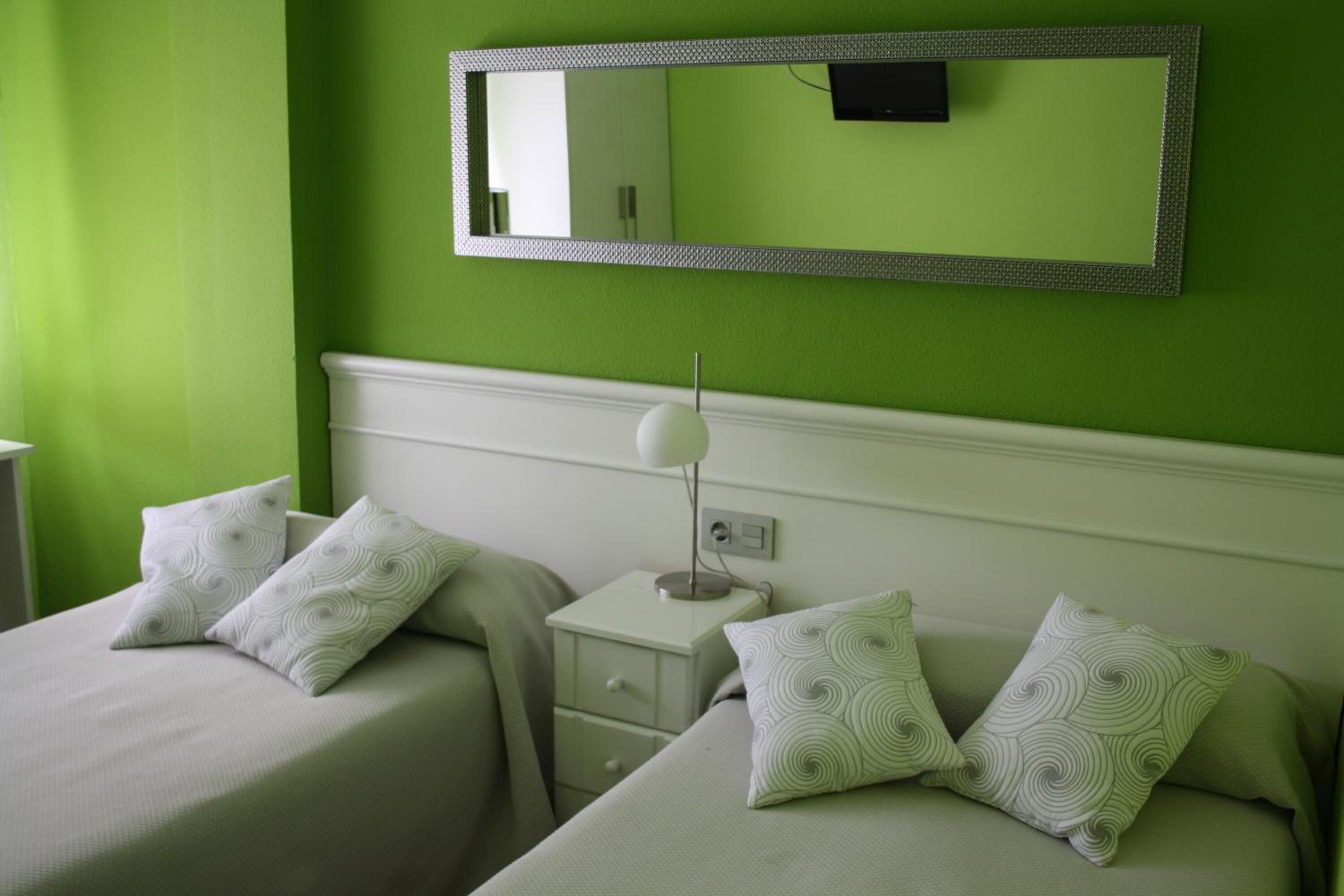 A room with two beds, each with a white bedspread and matching pillows, a bedside table with a lamp, and a large mirror on the wall.
