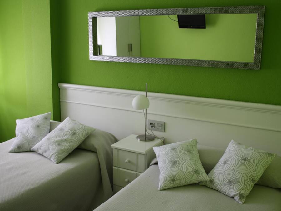 A room with two beds, each with a white bedspread and matching pillows, a bedside table with a lamp, and a large mirror on the wall.