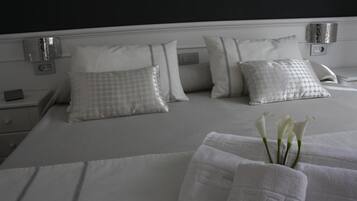 Una cama bien tendida con sábanas blancas y almohadas estampadas, una mesita de noche con lámpara, y un aplique de pared.
