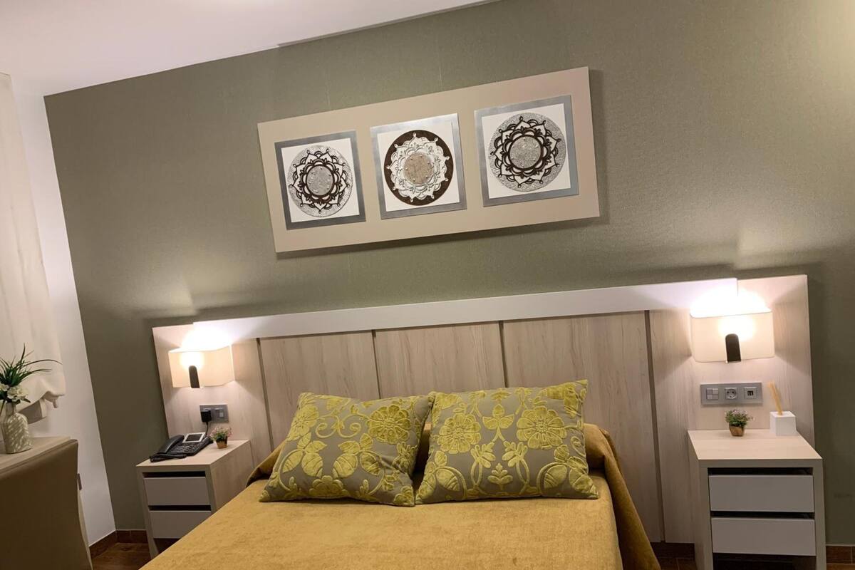 A bedroom with a bed, two bedside tables, a nightstand, a wall-mounted lamp, and three framed artworks above the bed.