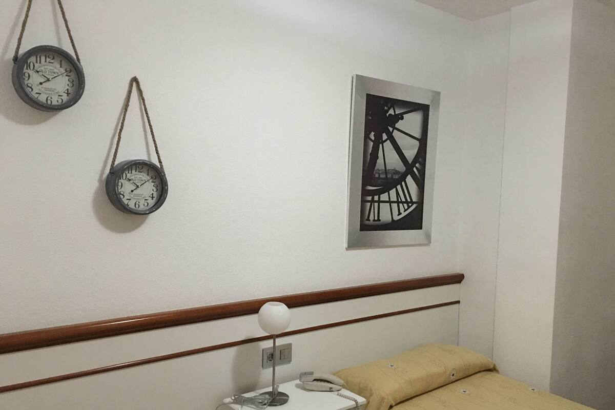 A bedroom with a bed, bedside table, lamp, two clocks, and a framed picture on the wall.