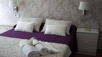 Una cama bien hecha con un edredón púrpura y almohadas blancas estampadas, una mesita de noche blanca con una lámpara y una toalla blanca doblada sobre la cama.