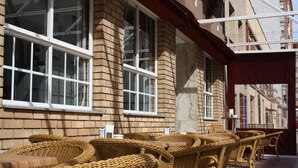 A row of wicker chairs and tables outside a building with large windows and a brick facade.