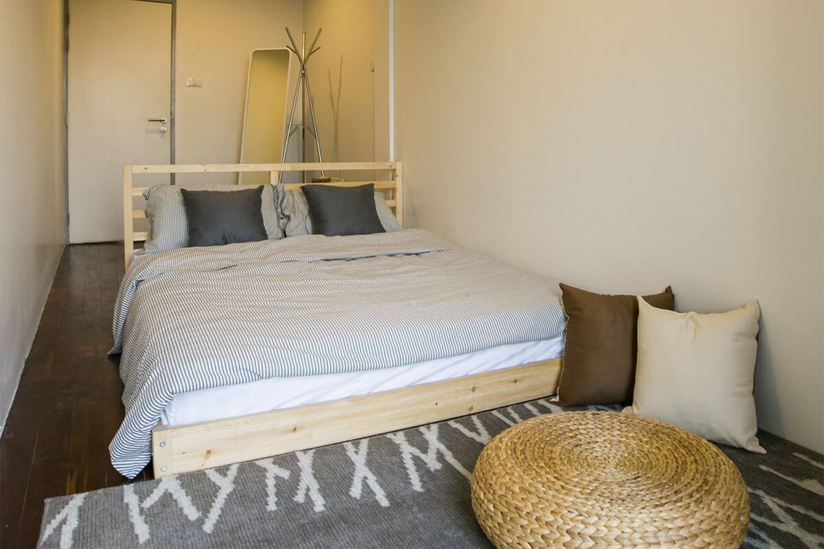 Un dormitorio con cama, almohadas, un reposapiés tejido, un espejo y una lámpara colgante.