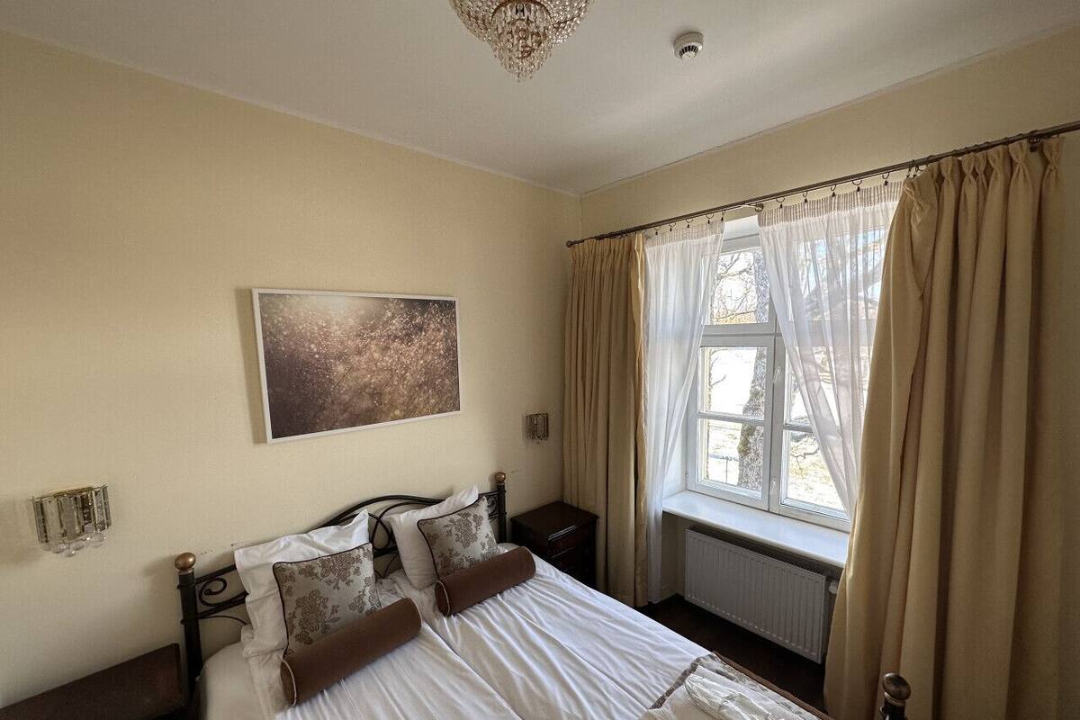 A bedroom with a bed, a nightstand, a radiator, a window with curtains, and a chandelier.