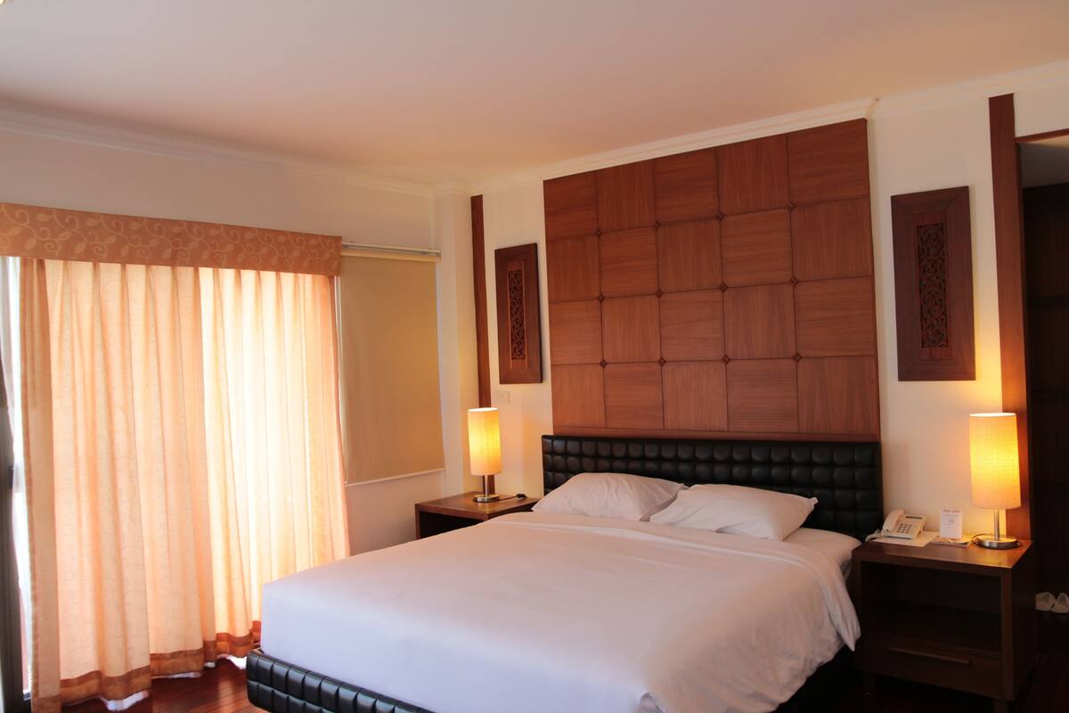 A hotel room with a large bed, wooden headboard, two bedside tables with lamps, and a window with curtains.