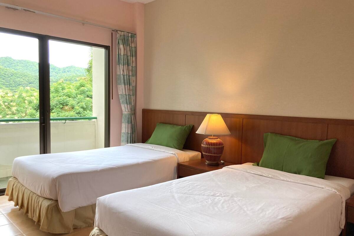 A hotel room with two beds, a nightstand with a lamp, and a view of greenery through a window.