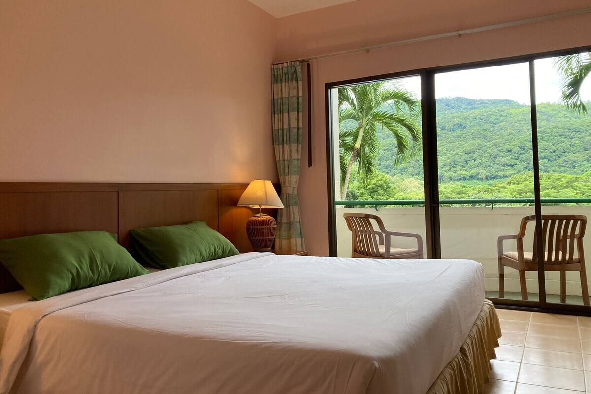 A hotel room with a large bed, a nightstand with a lamp, and a balcony with chairs.