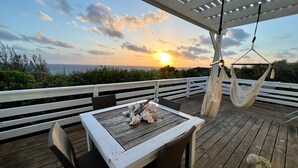 Eine Holzterrasse mit Tisch und Stühlen, einer Hängematte und Blick auf den Sonnenuntergang über dem Meer.