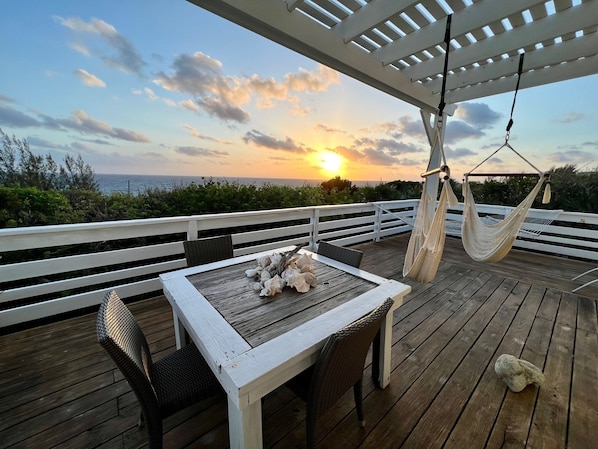 Una terraza de madera con una mesa y sillas, una hamaca colgante y vistas de la puesta de sol sobre el océano.