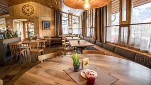 Een gezellig restaurantinterieur met houten tafels en banken, grote ramen en een klok aan de muur.