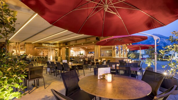 Een terras met ronde tafels en stoelen, rode parasols en een restaurant op de achtergrond.
