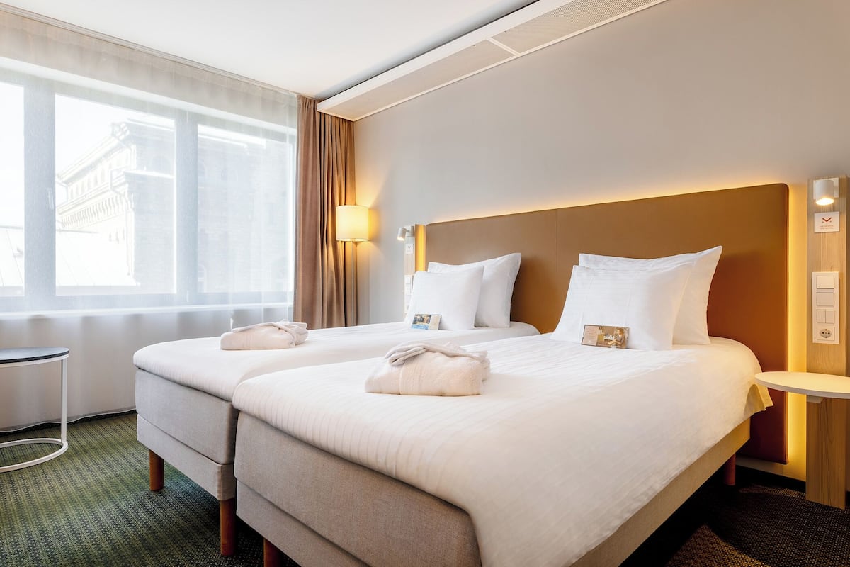 Habitación de hotel con dos camas, una mesita de noche, una lámpara y vista a un edificio desde la ventana.