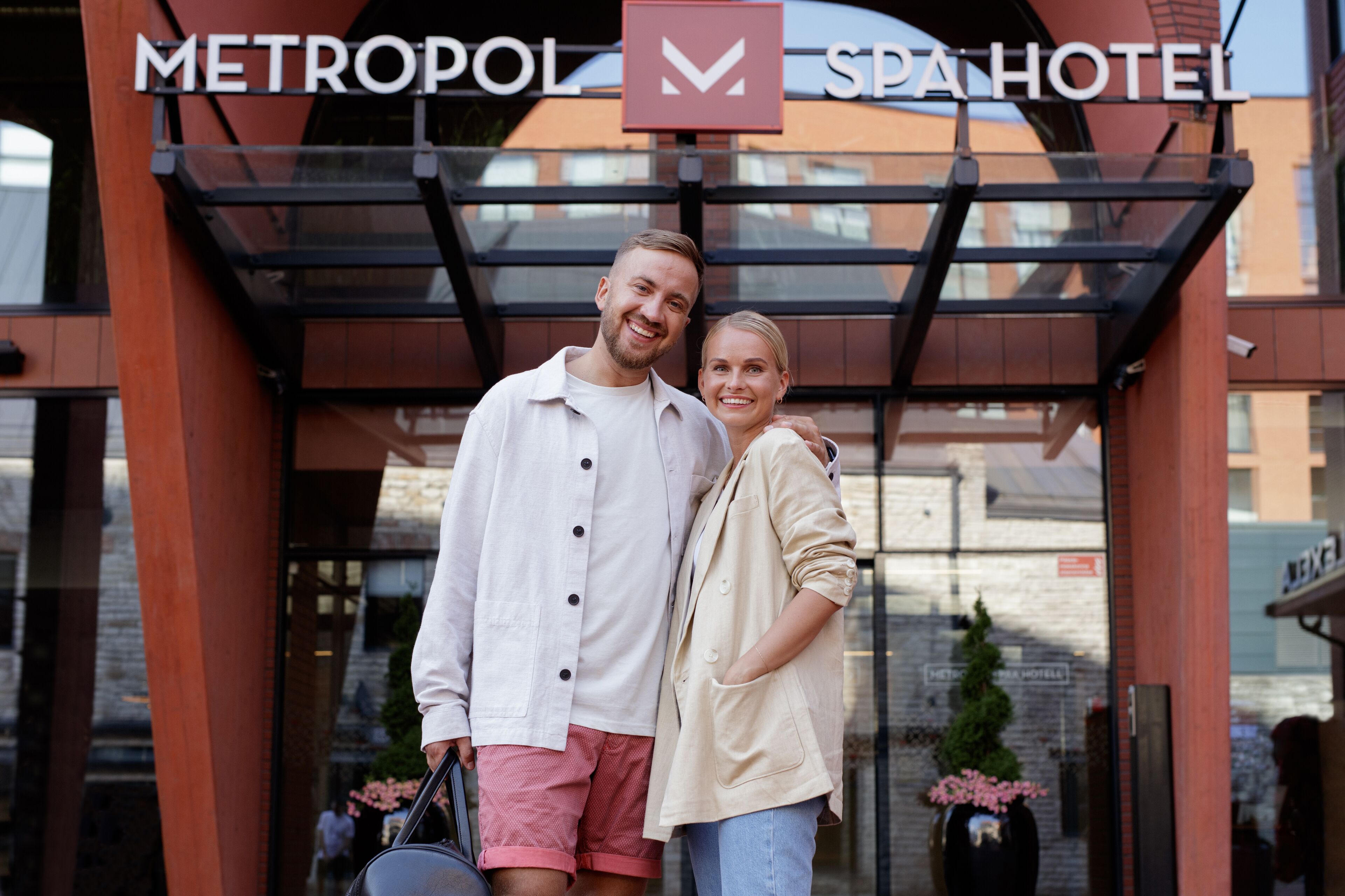 A couple standing in front of a hotel entrance with a glass canopy and a sign that reads "METROPOL SPA HOTEL".