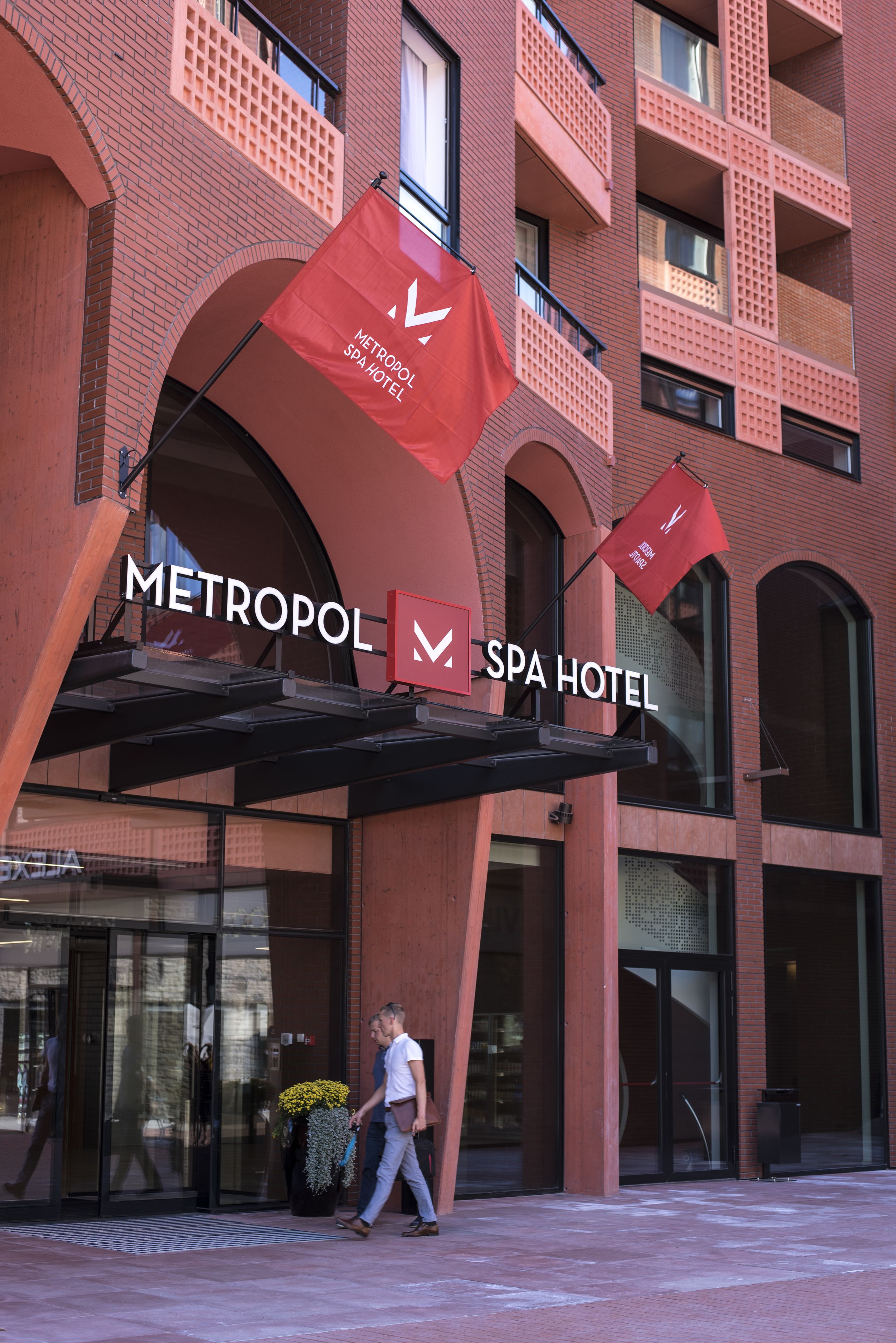 Exterior del Hotel Spa Metropol, con fachada de ladrillo moderno, amplio acceso en arco y banderas rojas que indican el nombre del hotel.