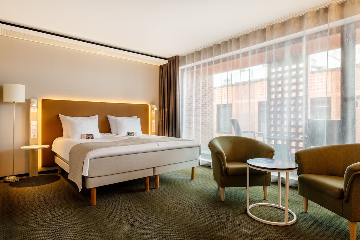 Una habitación de hotel con cama, dos sillones, una mesita y un ventanal con cortinas.