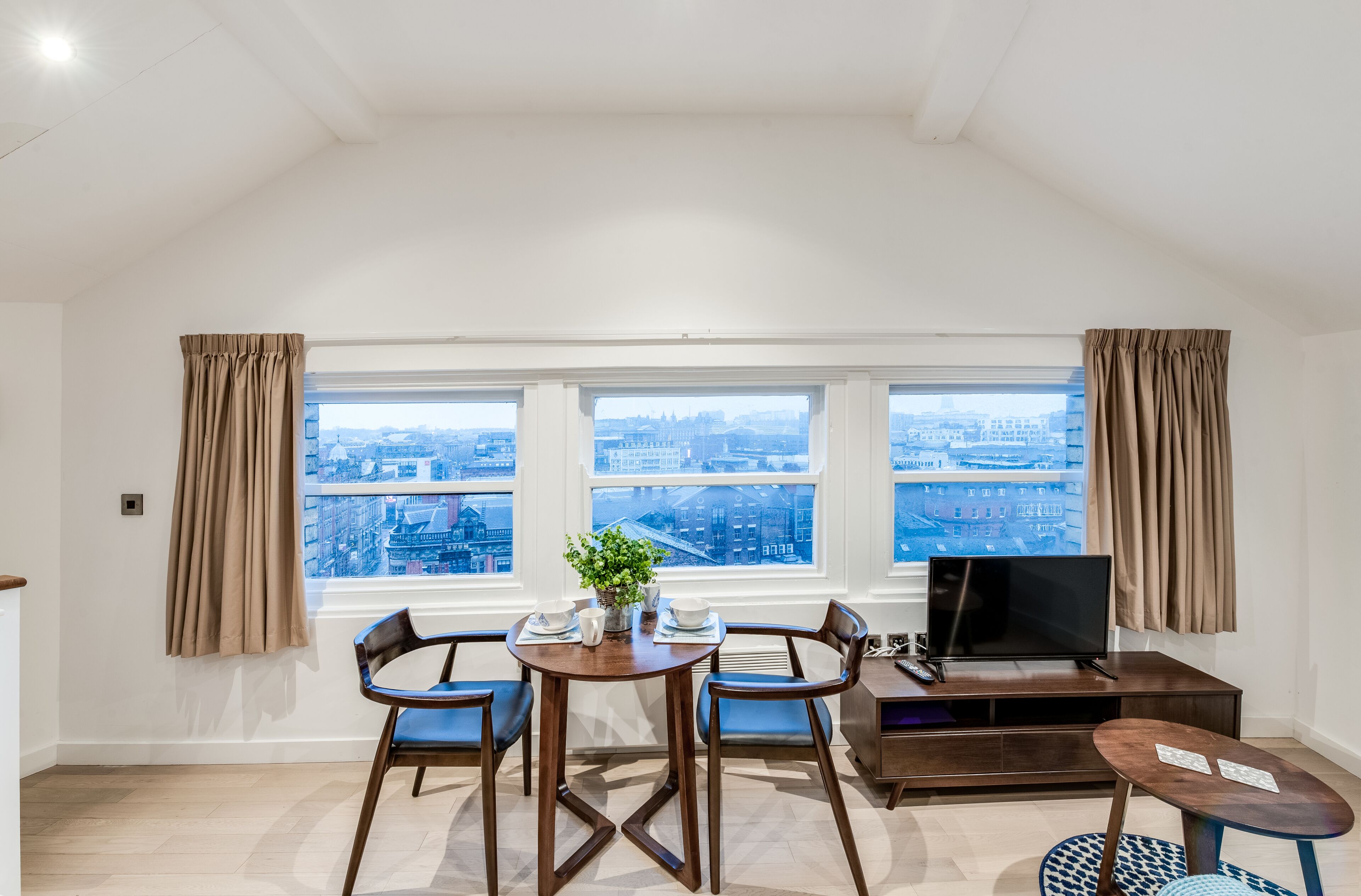 A modern living room with a dining area, large windows with city views, a flat-screen TV, and a small round table.