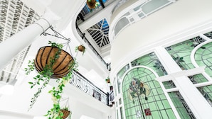 Un atrium à plusieurs niveaux, agrémenté de jardinières suspendues, d’une grande fenêtre en vitrail aux motifs végétaux et de colonnes blanches.