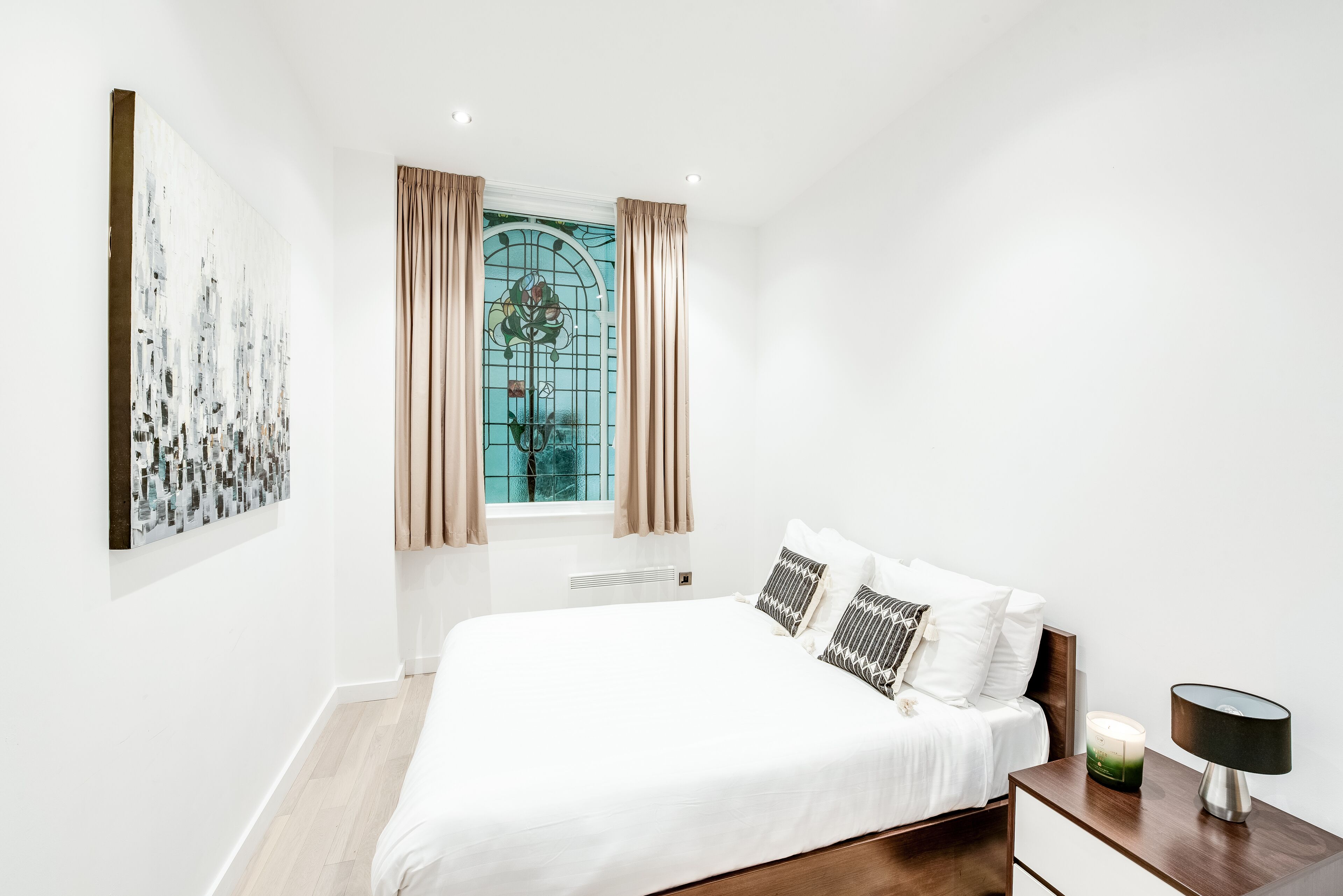 Un dormitorio con una cama grande, una mesita de noche, una lámpara de pared y un ventanal con cortinas.