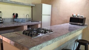 Eine Küche mit Gasherd, Mikrowelle und Kühlschrank. Zwei Hocker und ein Spülbecken mit Geschirrgestell sind vorhanden.