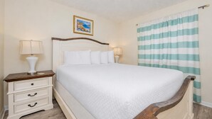 Una cama bien tendida con ropa blanca, un cabecero de madera, una mesita de noche con una lámpara y una cortina a rayas.