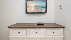 Un televisor fijado a la pared mostrando una escena de playa, un aparador de madera con cajones y un termostato pequeño también en la pared.