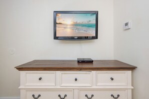 Un televisor fijado a la pared mostrando una escena de playa, un aparador de madera con cajones y un termostato pequeño también en la pared.