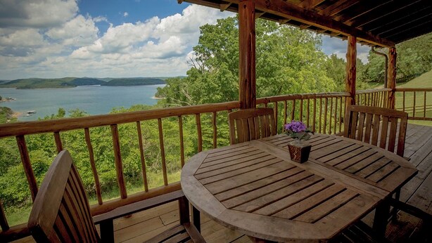 Um deck de madeira com mesa e cadeiras, com vista para um lago e colinas.