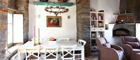Un comedor con una mesa y sillas blancas, una araña, una chimenea de piedra y una estantería.