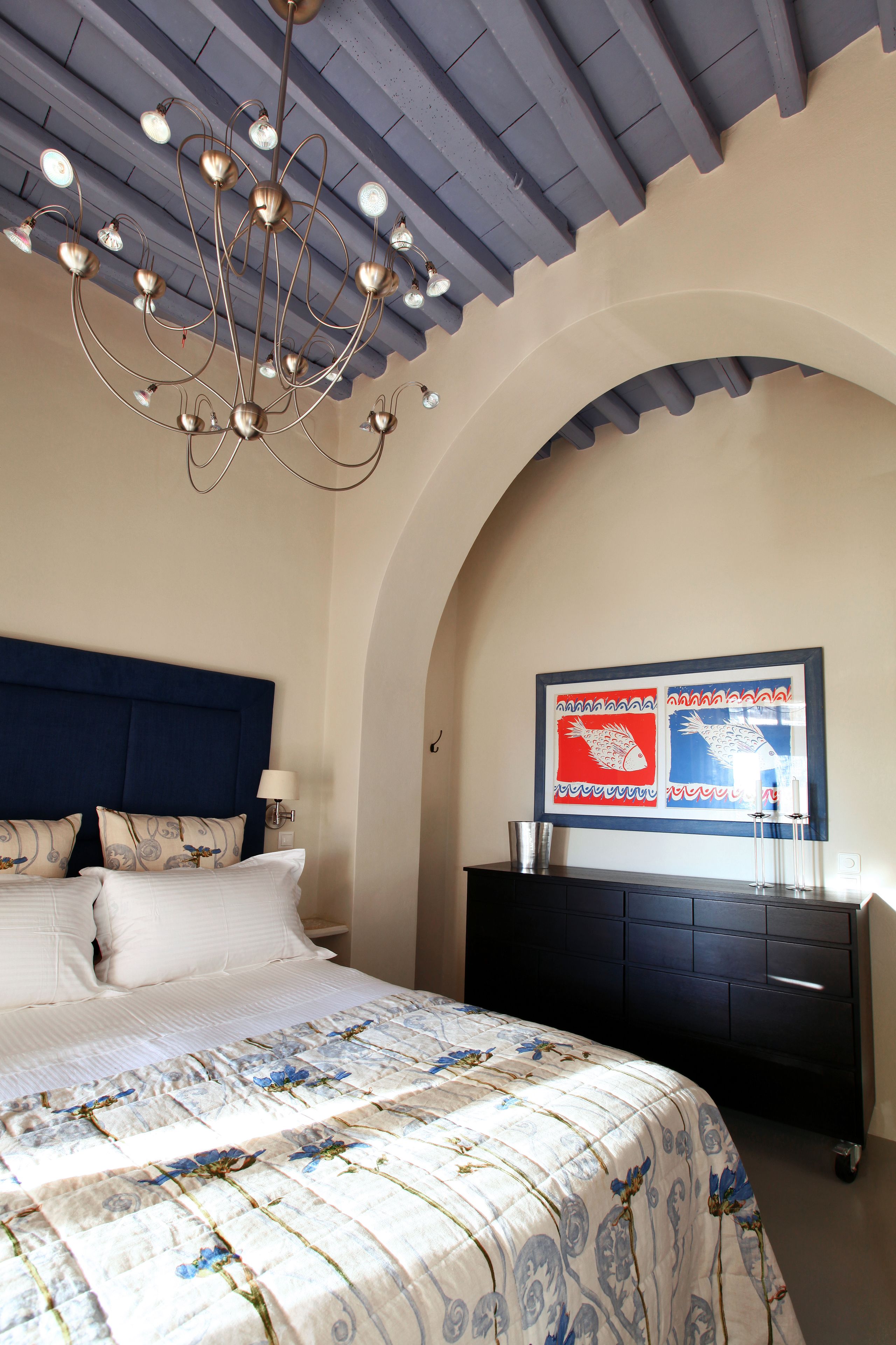 Ein Schlafzimmer mit einem großen Bett, einem dunklen Kopfteil, einer Kronleuchter, einem an der Wand befestigten Kunstwerk und einer dunklen Kommode.