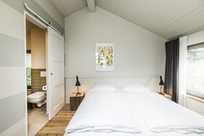 A bedroom with a bed, bedside tables, a nightstand, a window with curtains, a door leading to a bathroom, and a wall-mounted artwork.