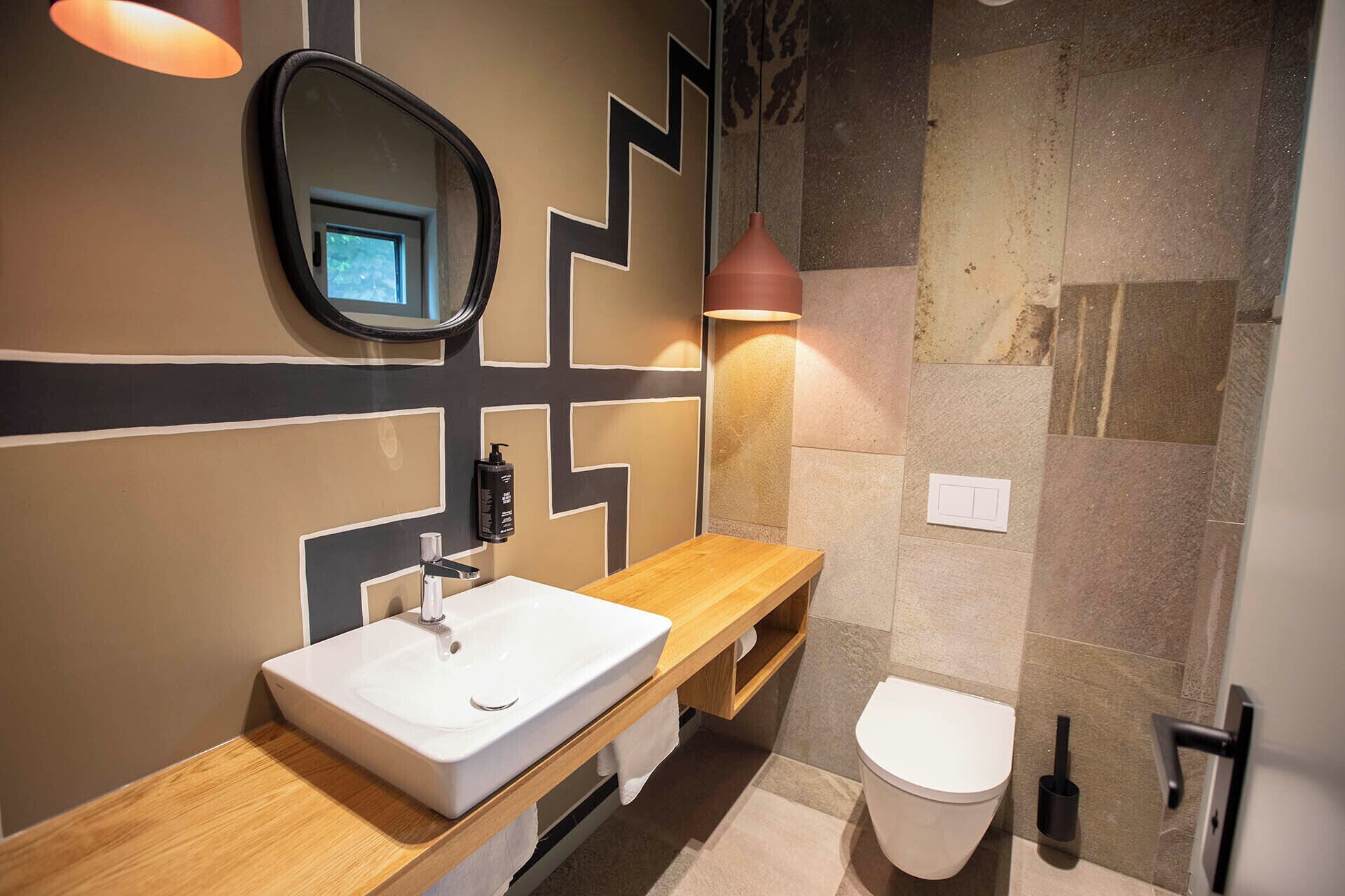 A modern bathroom with a white sink, wooden vanity, a mirror, a wall-mounted light, and a toilet.