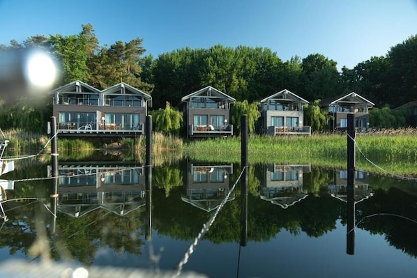 A row of modern houses with balconies, reflecting in calm water, surrounded by greenery.