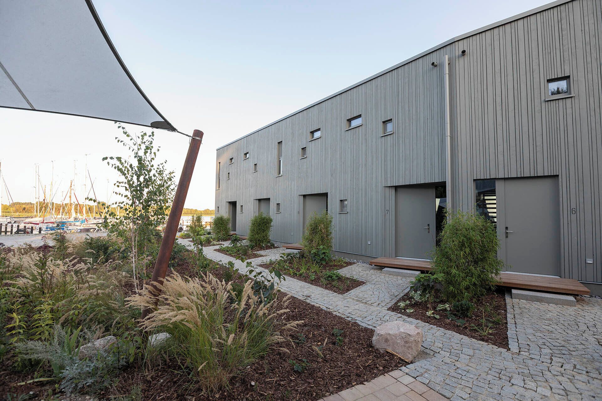 Een modern gebouw met een plat dak, meerdere ramen en een geplaveid pad met plantenbakken.