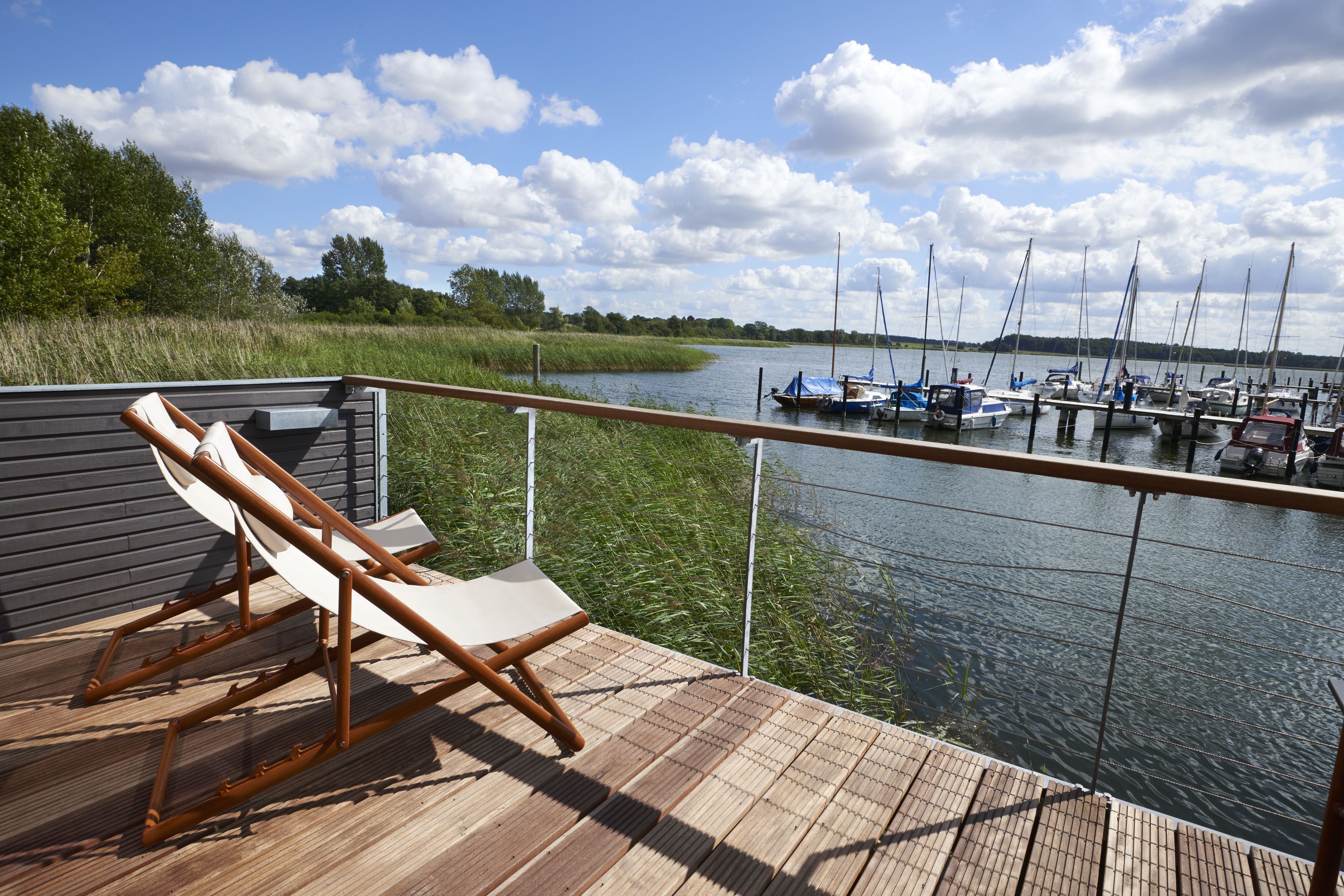 Eine Holzterrasse mit zwei Liegestühlen mit Blick auf ruhiges Wasser und mehreren dort liegenden Booten.