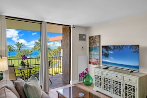 A living room with a television, a sofa, a coffee table, and a view of the ocean through a sliding glass door. - Penthouse Oceanview Vistas (Kihei, Maui)