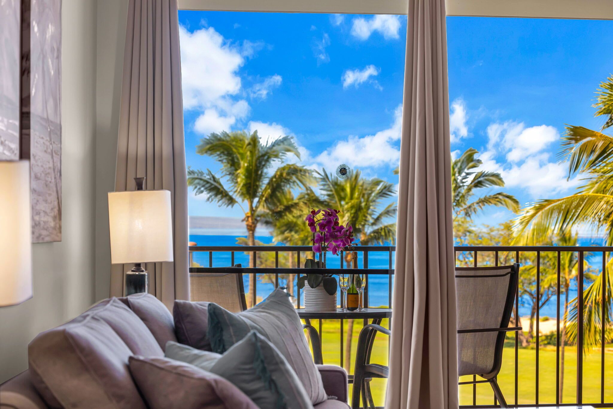 Ein Zimmer mit Blick auf Palmen und das Meer, ein Balkon mit Tisch für zwei Personen und eine Couch mit Kissen.