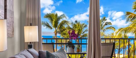 Ein Zimmer mit Blick auf Palmen und das Meer, ein Balkon mit Tisch für zwei Personen und eine Couch mit Kissen.