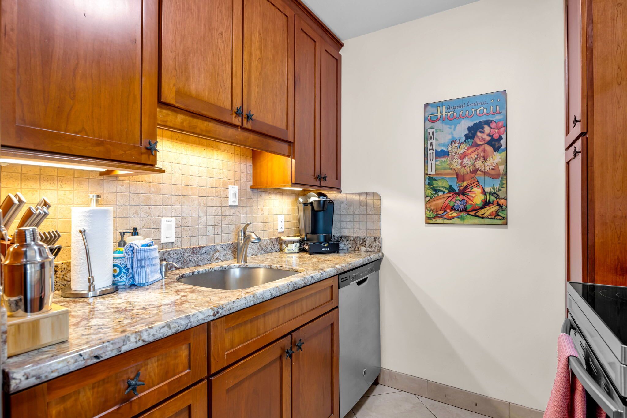 Eine Küche mit Holzschränken, einer Marmorarbeitsplatte, einer Kaffeemaschine und einem Poster mit hawaiianischem Dekor.