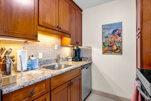 A kitchen with wooden cabinets, a marble countertop, a coffee maker, and a Hawaiian-themed poster. - Penthouse Oceanview Vistas (Kihei, Maui)