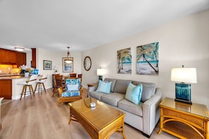 A living room with a sofa, coffee table, and dining area with chairs. There are paintings on the wall and a lamp on a side table. - Penthouse Oceanview Vistas (Kihei, Maui)