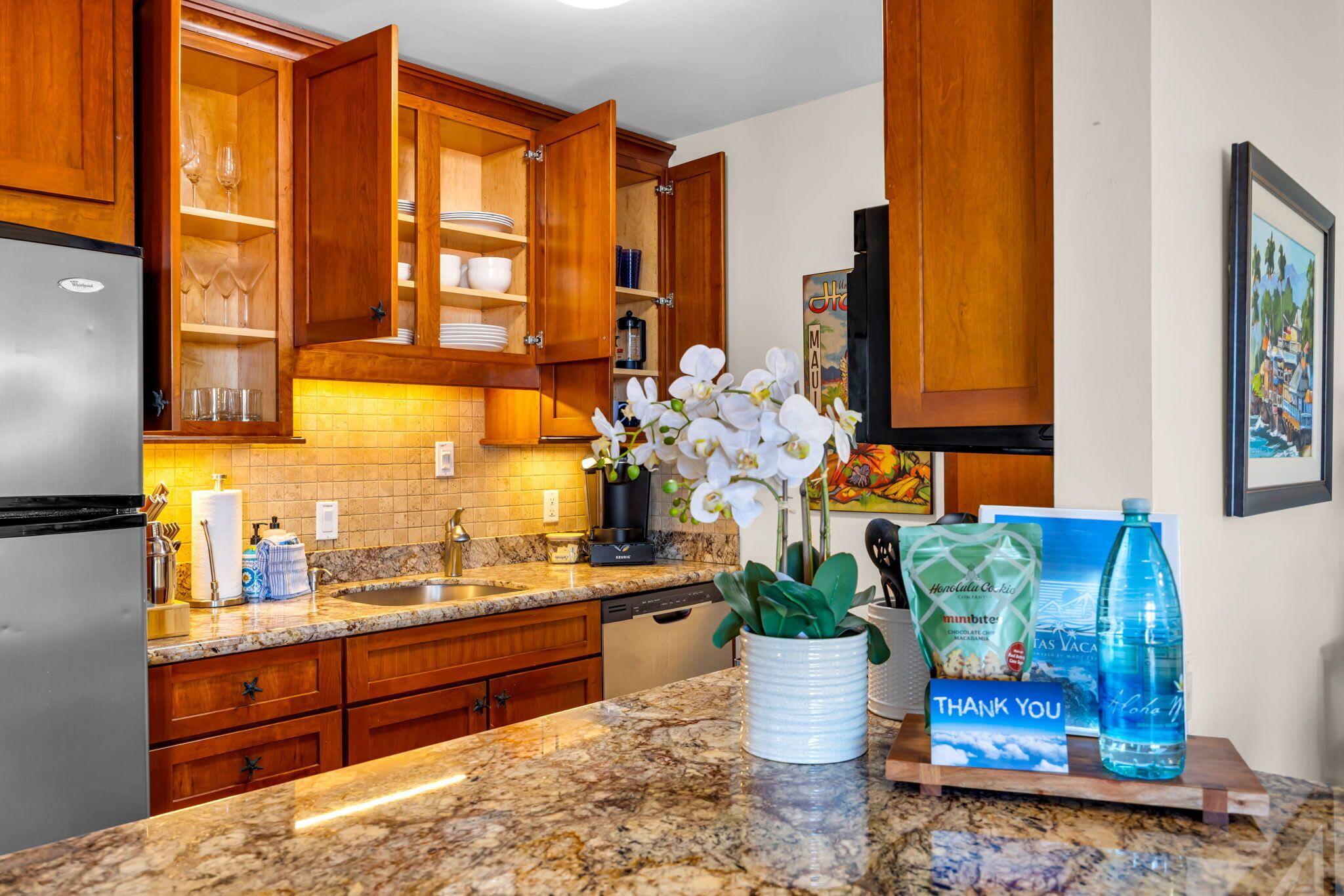 Una cocina con armarios de madera, encimera de granito, refrigerador y un florero con flores blancas.