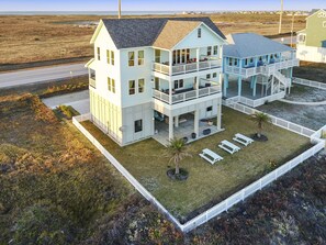A two-story beachfront house with a balcony, a covered patio, and a fenced lawn area.